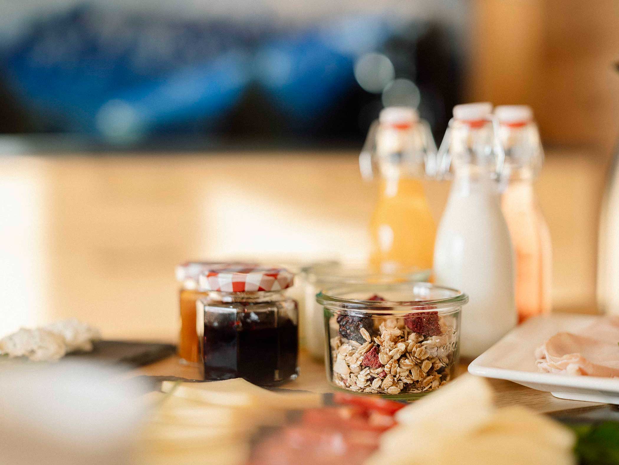 Frühstücksartikel auf einem Tisch: Müsli im Glas, Marmelade, Getränke in Flaschen und eine Platte mit Käse und Wurst. - Langfennhof