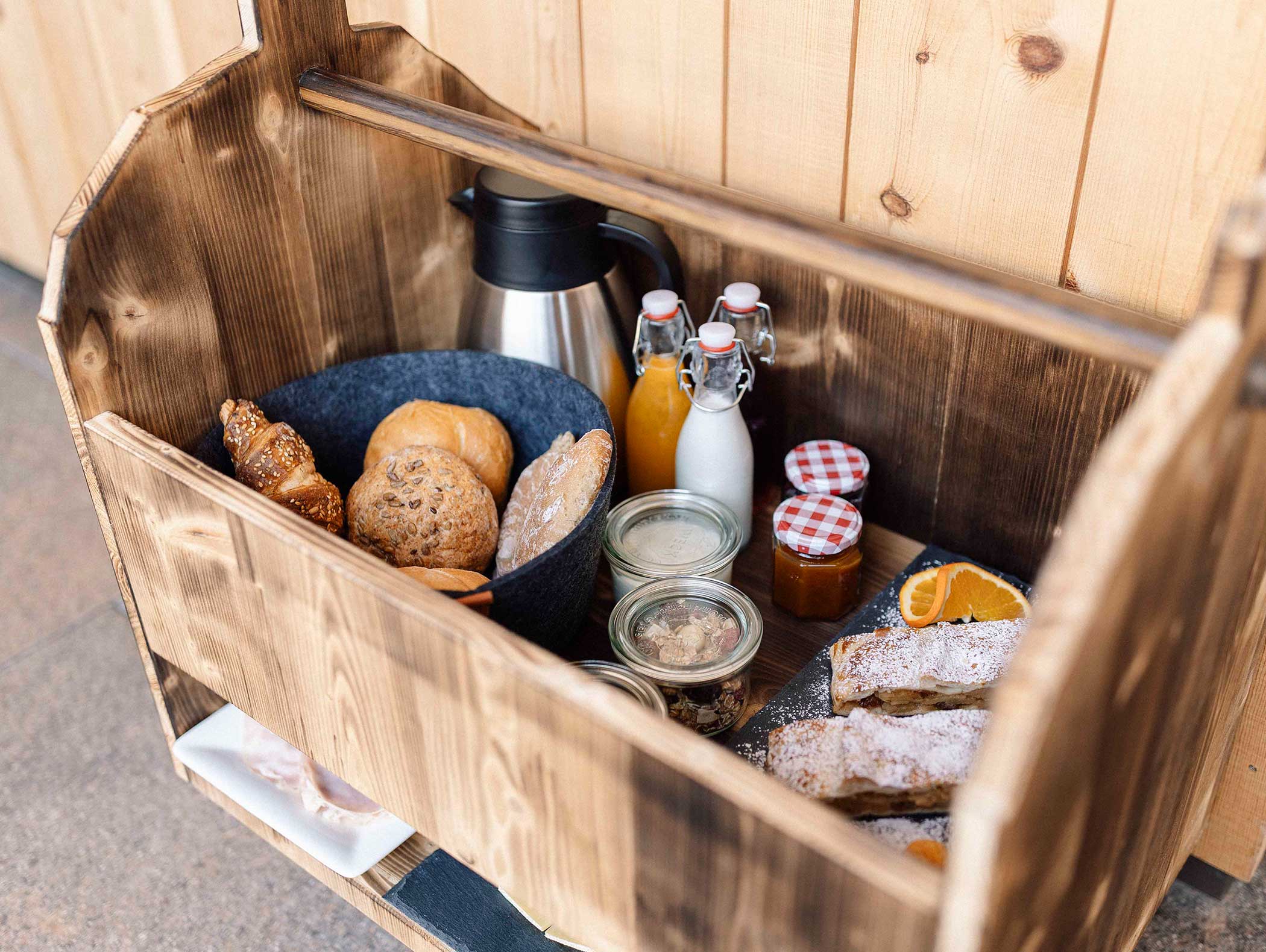 Holzkiste mit Brot, Gebäck, einer Thermoskanne, Saftflaschen, Marmeladengläsern und einer Schale mit gemischten Nüssen. - Langfennhof