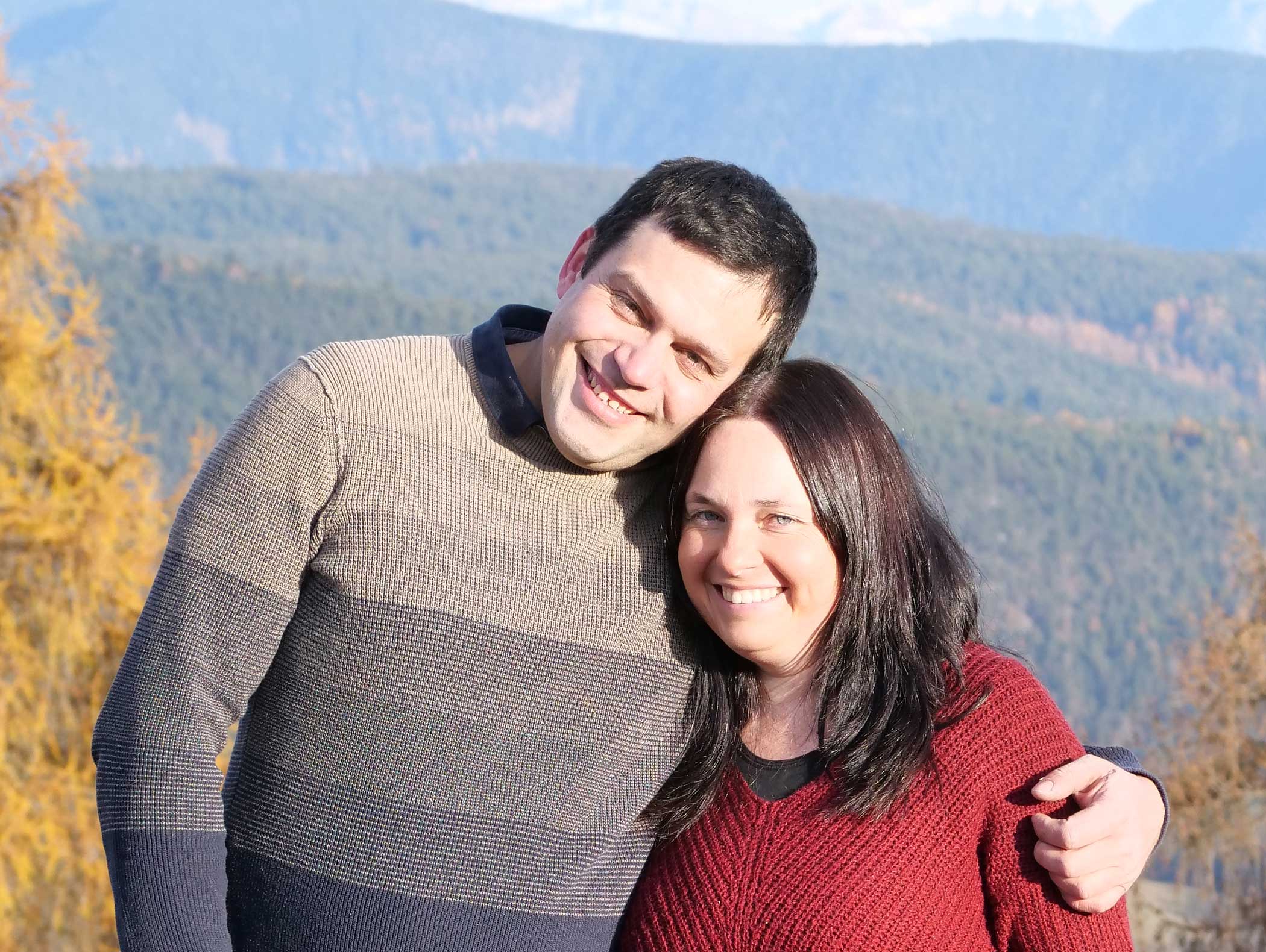 Ein Mann und eine Frau lächeln zusammen im Freien, mit Bergen und Bäumen im Hintergrund. - Langfennhof