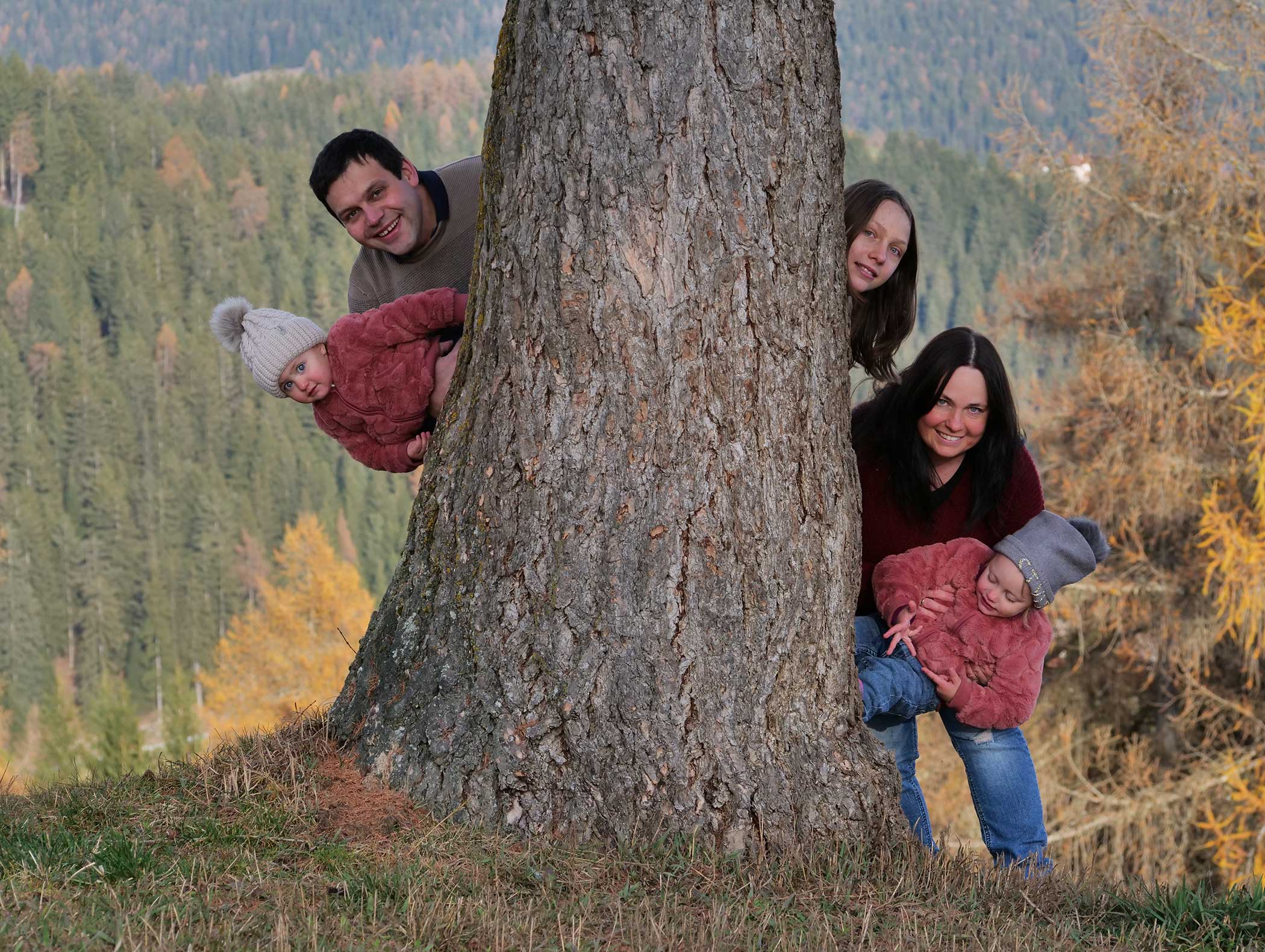 Eine fünfköpfige Familie lächelt, während sie hinter einem großen Baum in einem Waldgebiet hervorschaut. - Langfennhof
