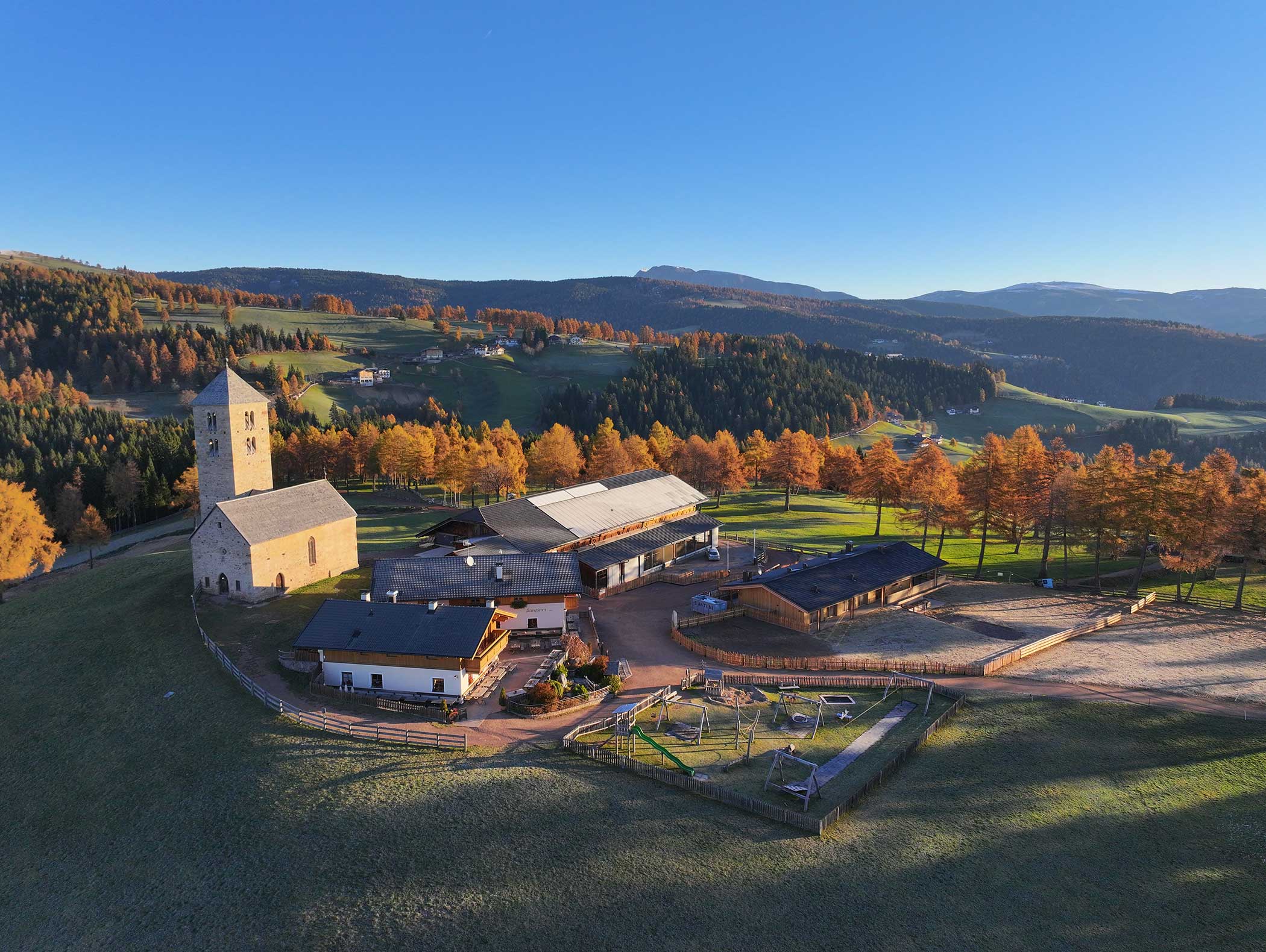Luftaufnahme eines ländlichen Bauernhofs und einer Kirche, umgeben von herbstlichen Bäumen und sanften Hügeln unter einem klaren blauen Himmel. - Langfennhof