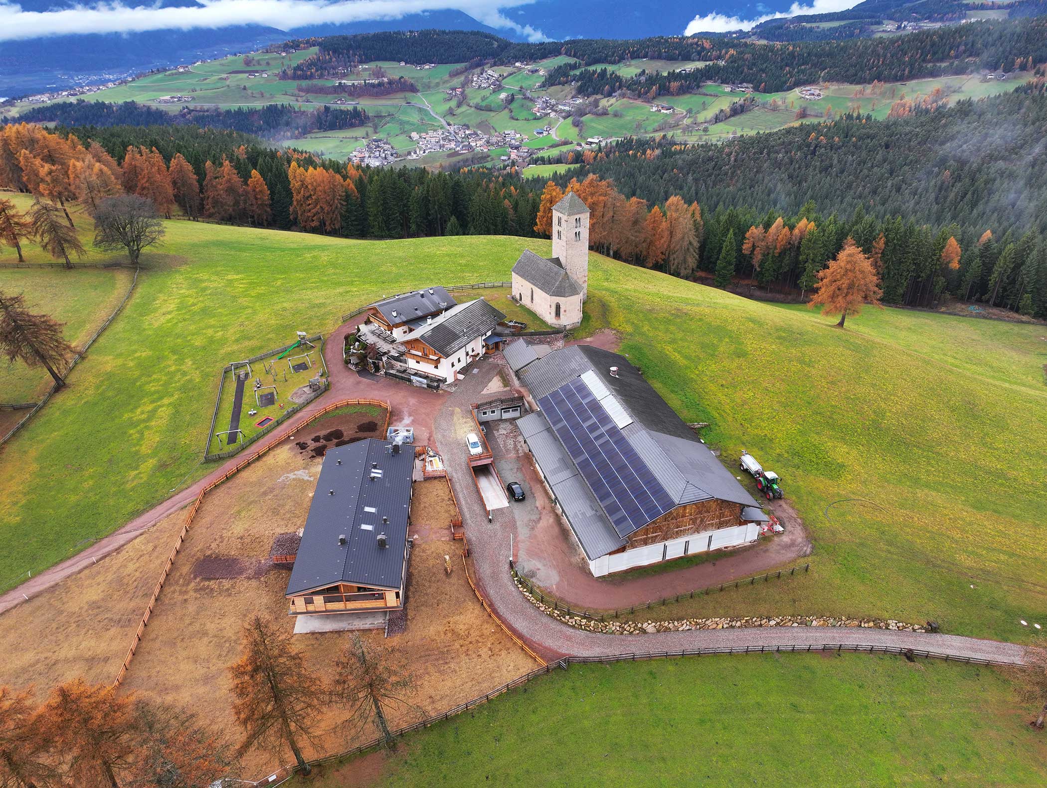 Luftaufnahme eines ländlichen Bauernhofs mit Sonnenkollektoren, Gebäuden und einer Kirche auf einer grünen Anhöhe. - Langfennhof