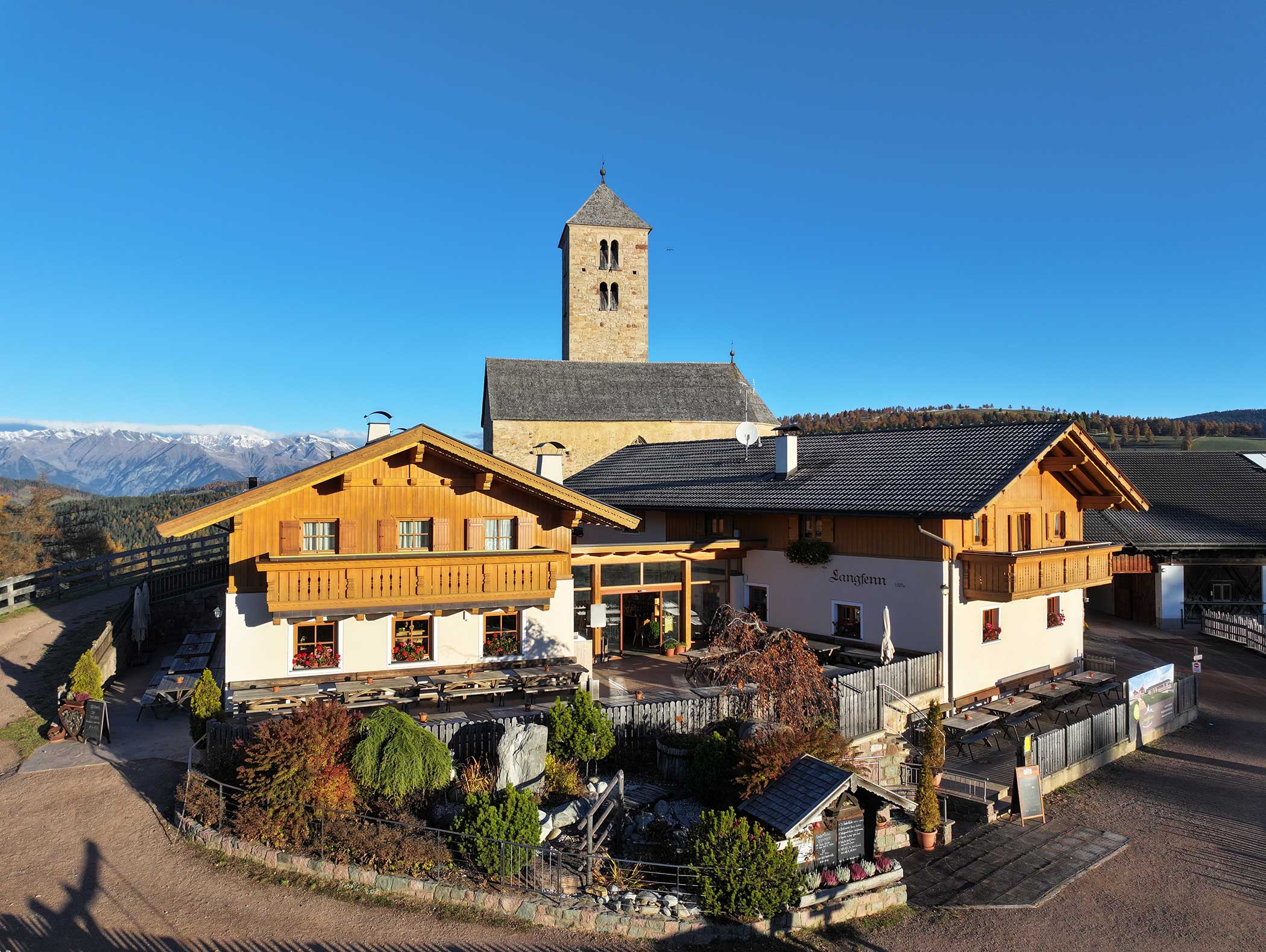 Charmantes Alpengasthaus mit Holzbalkonen und steinernem Kirchturm in einem Bergdorf unter strahlend blauem Himmel. - Langfennhof