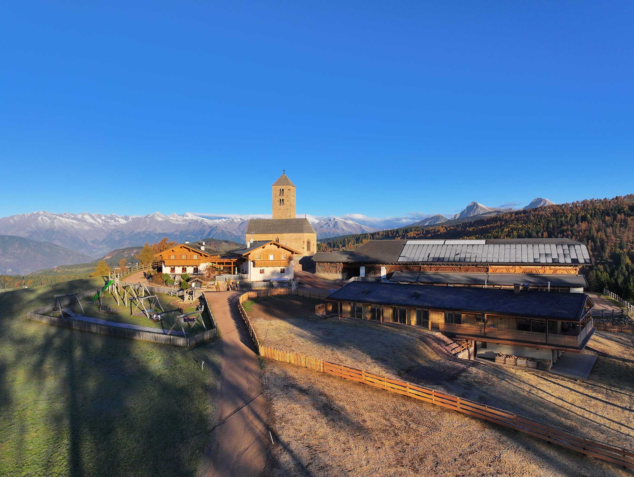 Auf einem Hügel gelegene Bauernhäuser mit einem Kirchturm, umgeben von Bergen unter einem klaren blauen Himmel. - Langfennhof