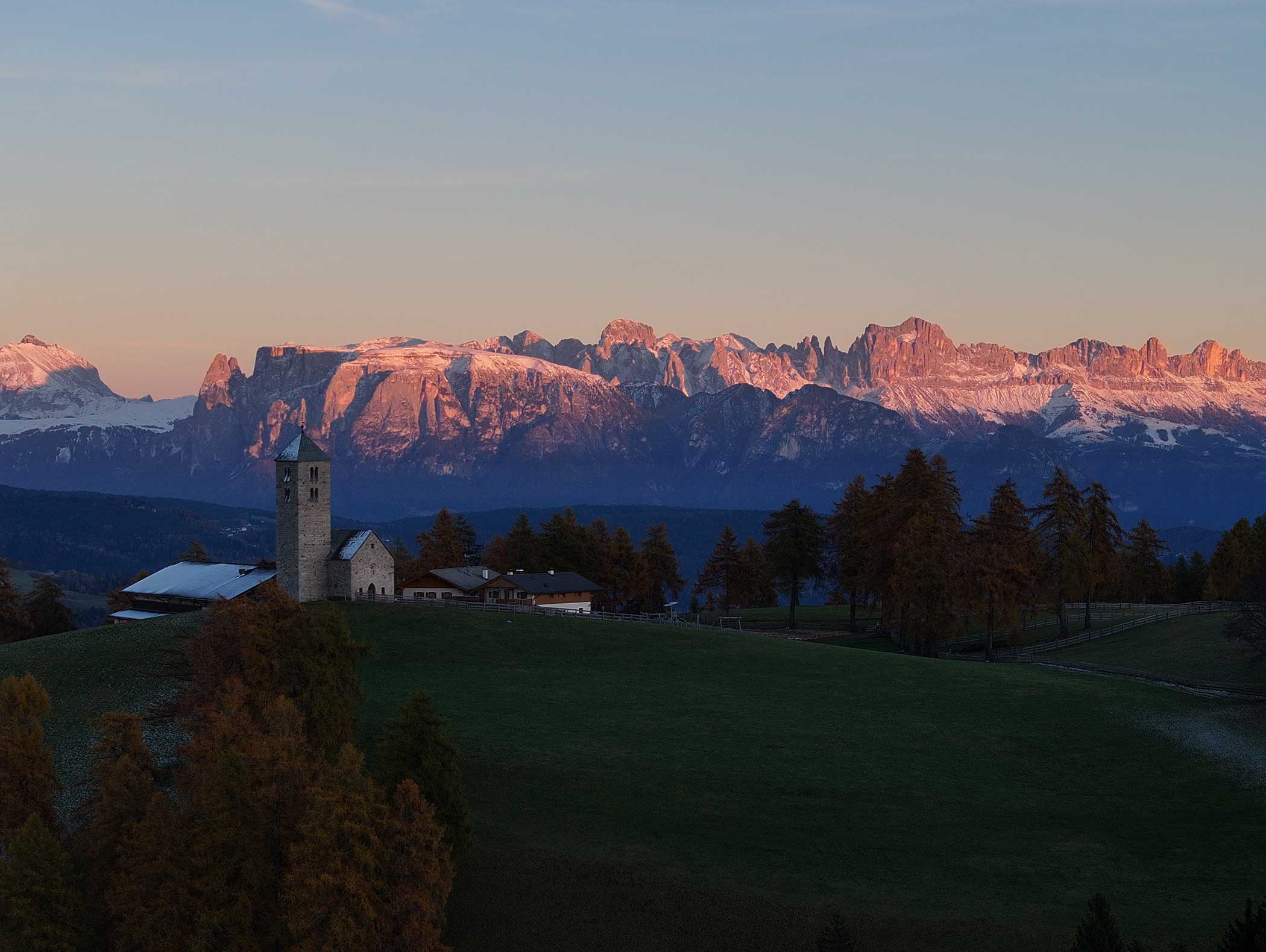 Eine Kirche und ein Dorf stehen vor Bergen, die vom rosafarbenen Licht des Sonnenuntergangs angestrahlt werden, mit Bäumen und grünen Feldern im Vordergrund. - Langfennhof