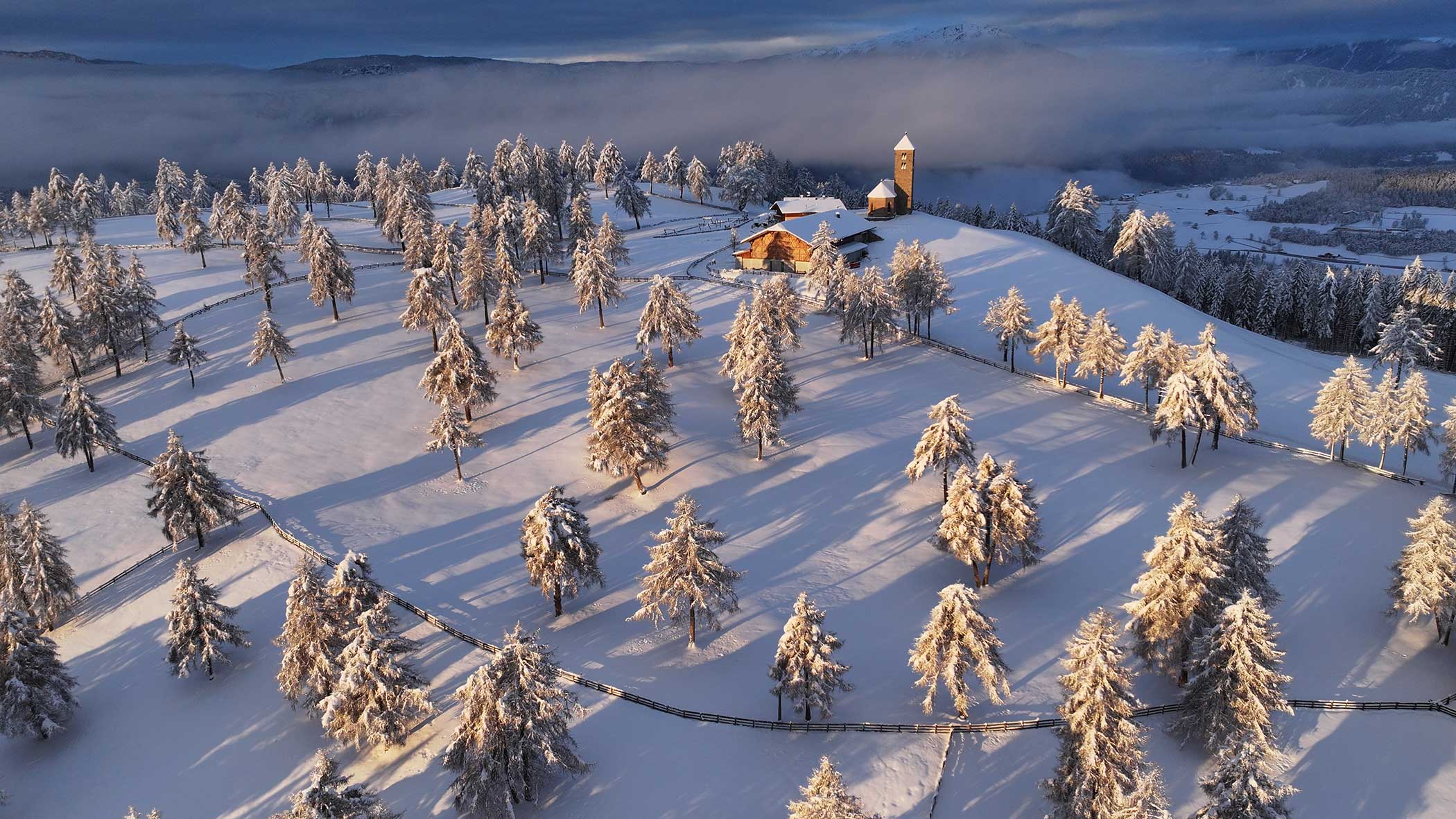 Luftaufnahme eines verschneiten Hügels mit verstreuten Bäumen, einer Kirche und Häusern unter einem dramatischen Himmel. - Langfennhof
