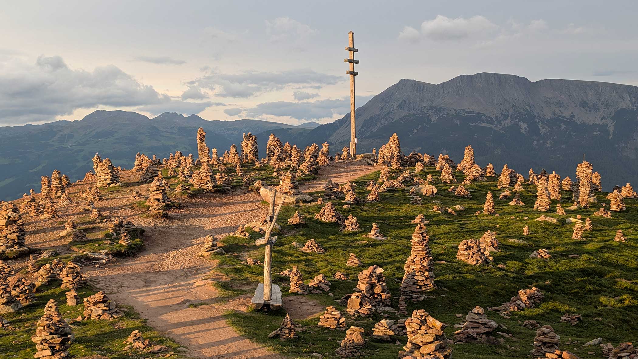 Eine Hügelkuppe mit kleinen Steinhaufen, unbefestigten Wegen und einem hohen Holzpfahl, im Hintergrund die Berge. - Langfennhof