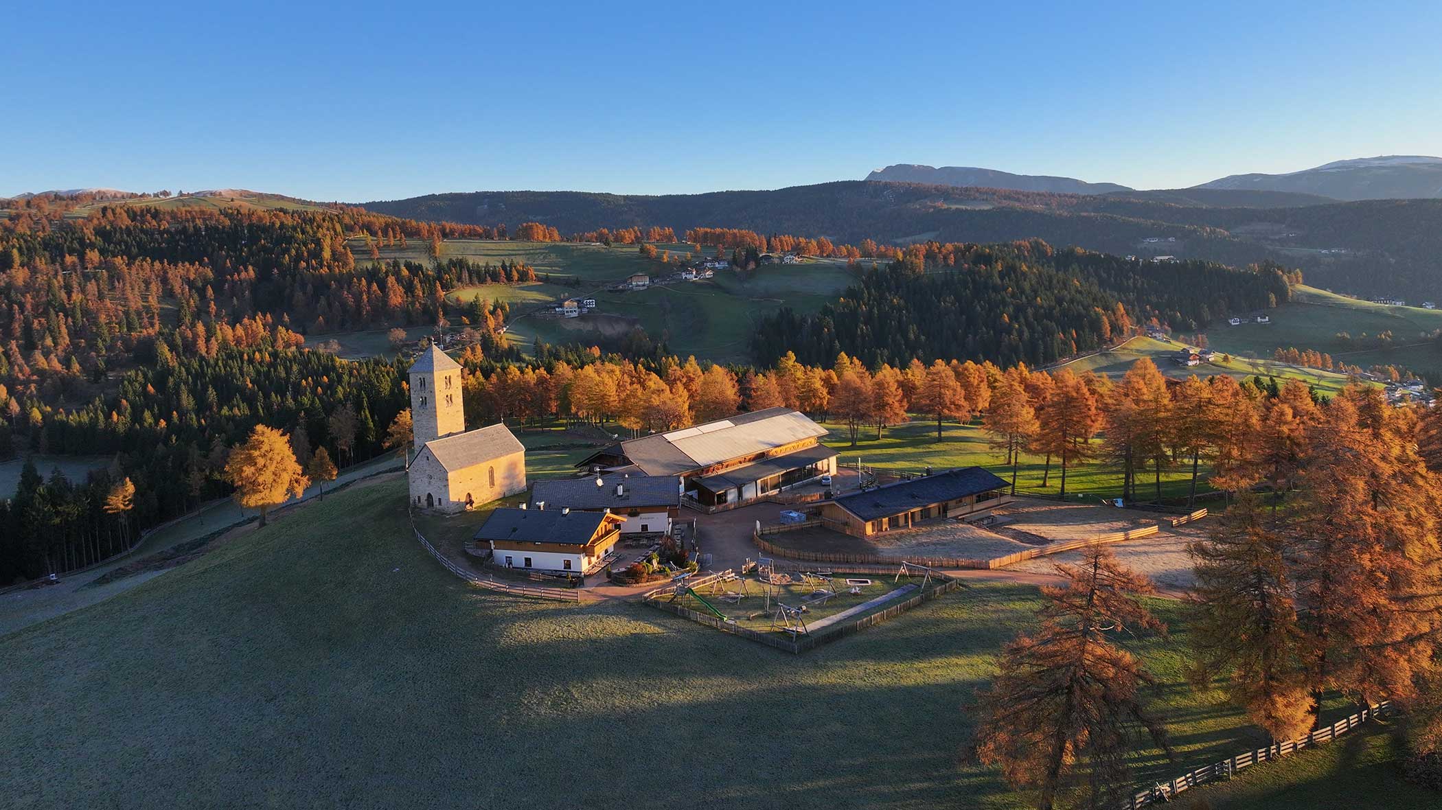 Luftaufnahme eines Bauernhofs und einer Kirche auf einem Hügel, umgeben von Herbstbäumen und fernen Bergen. - Langfennhof