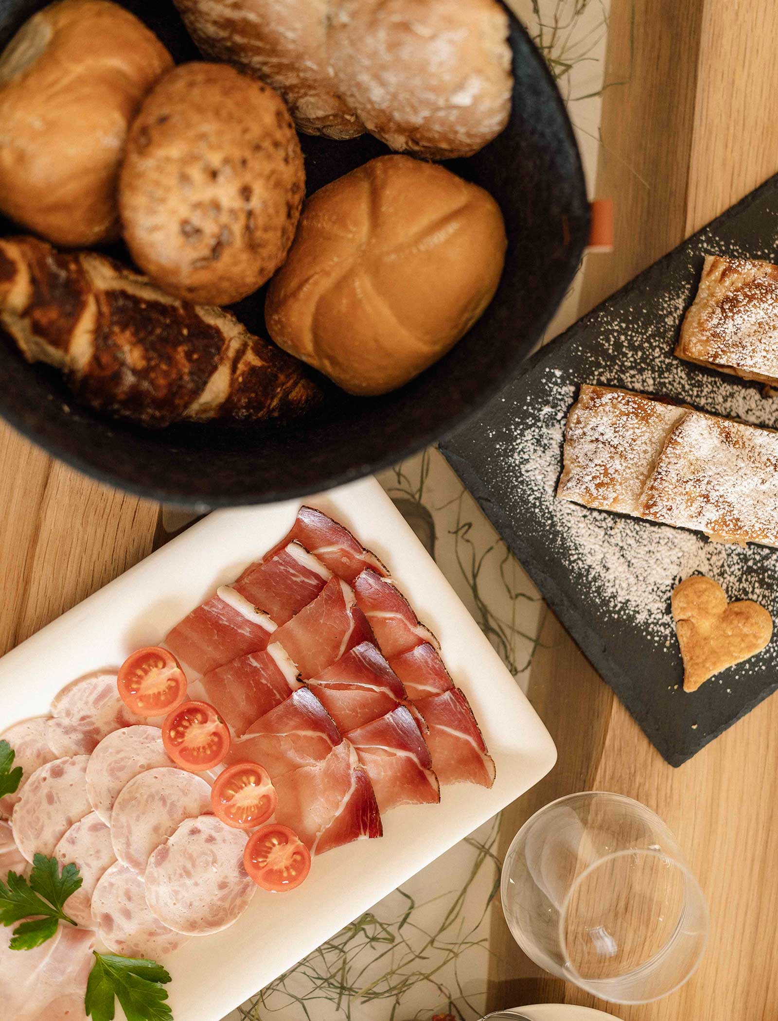 Ein Frühstücksaufstrich mit verschiedenen Brötchen, Aufschnitt, Kirschtomaten und mit Puderzucker bestäubtem Gebäck. - Langfennhof