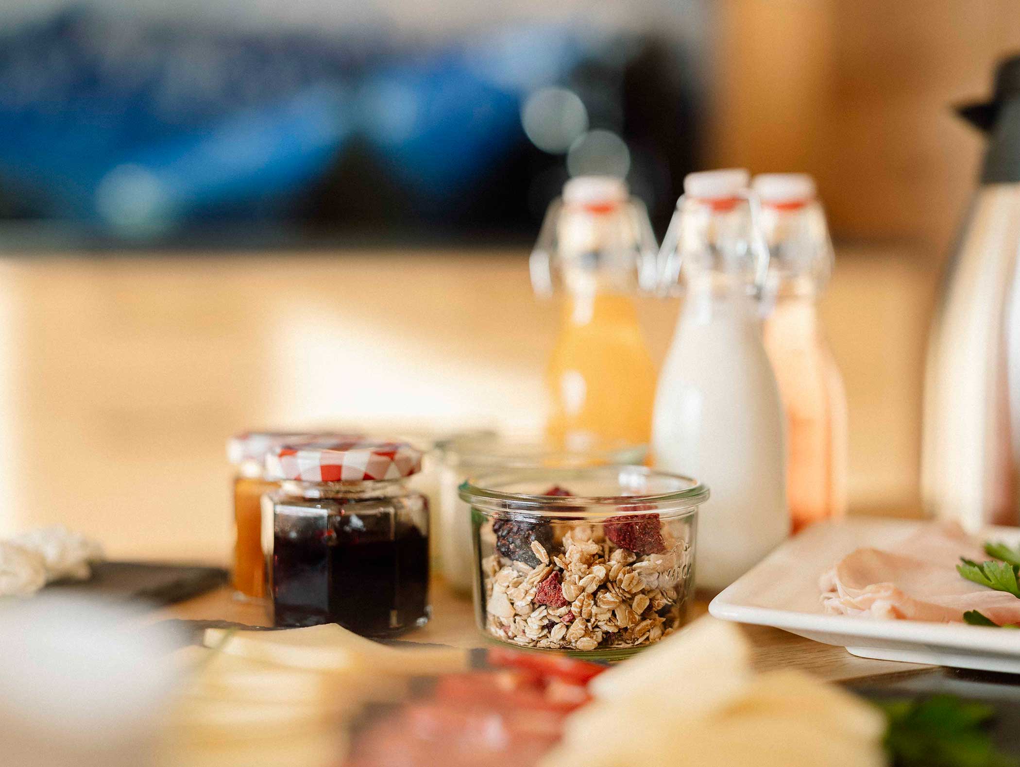 Gläser mit Marmelade und Müsli, Flaschen mit Saft und Milch und Aufschnitt auf einem Frühstückstisch. - Langfennhof