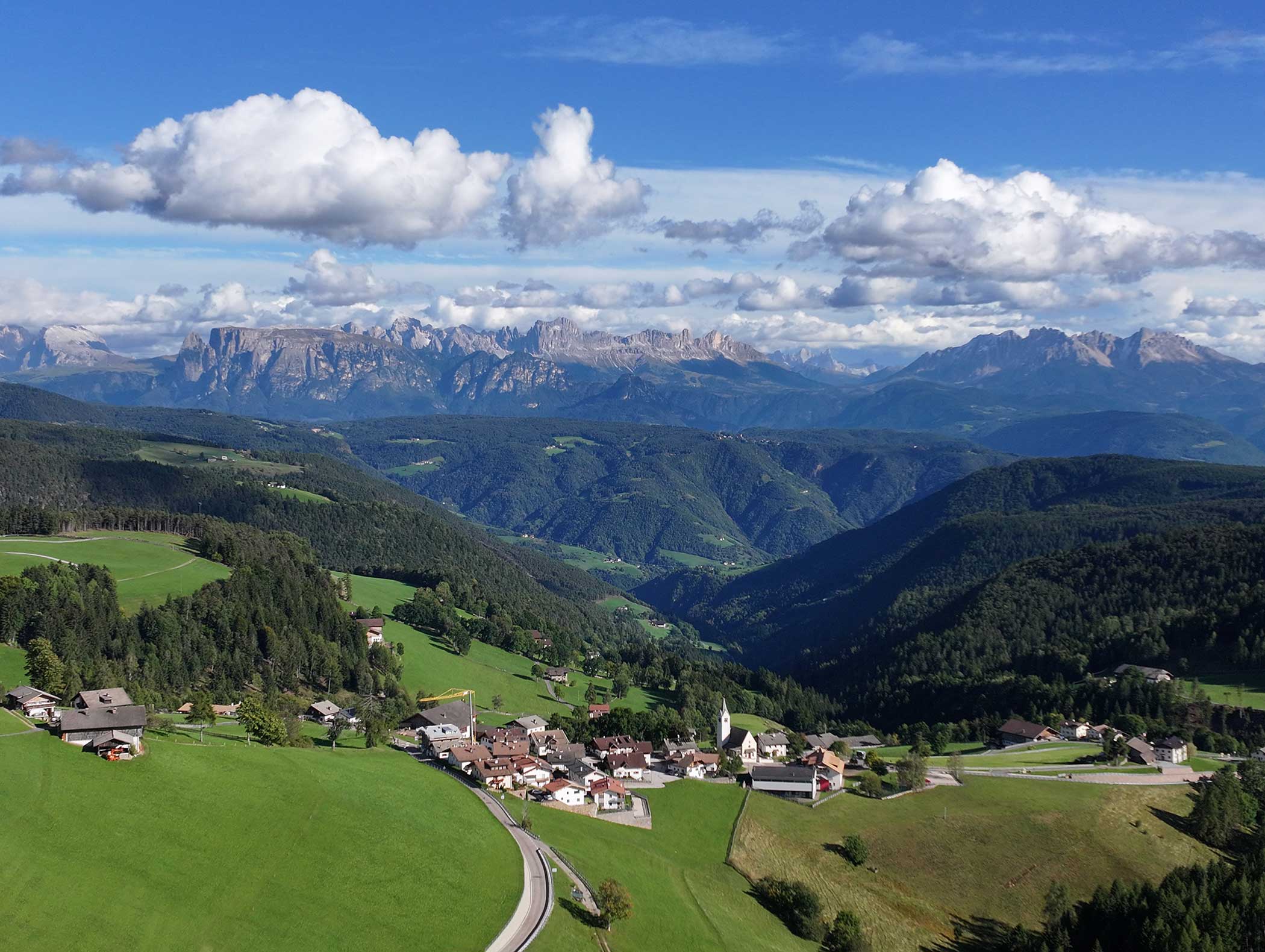 Bergdorf mit verstreuten Häusern, grünen Hügeln, Wäldern und entfernten Gipfeln unter einem teilweise bewölkten Himmel. - Langfennhof