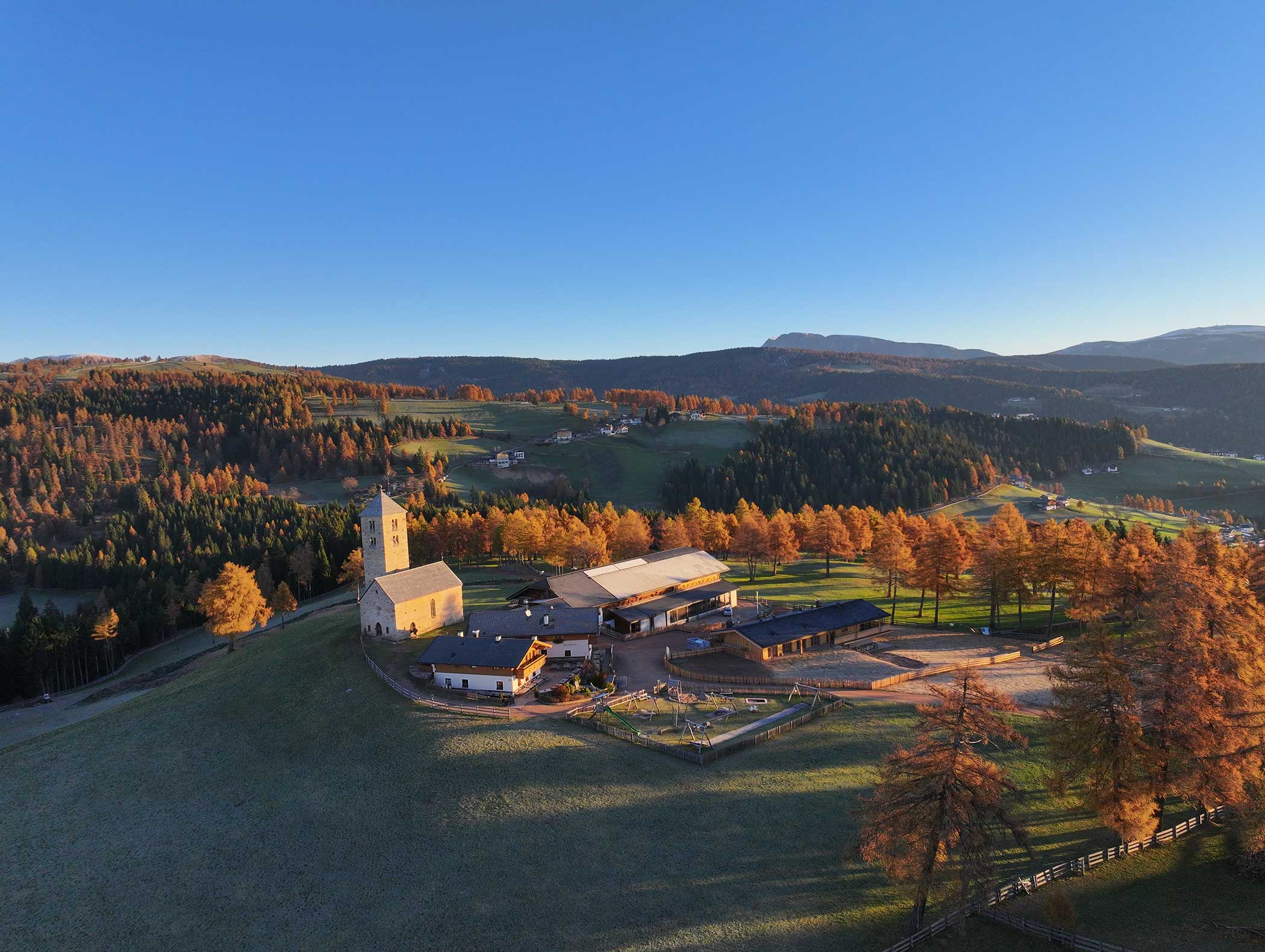 Luftaufnahme eines ländlichen Bauernhofs und einer Kirche, umgeben von herbstlichen Bäumen und sanften Hügeln unter einem klaren blauen Himmel. - Langfennhof