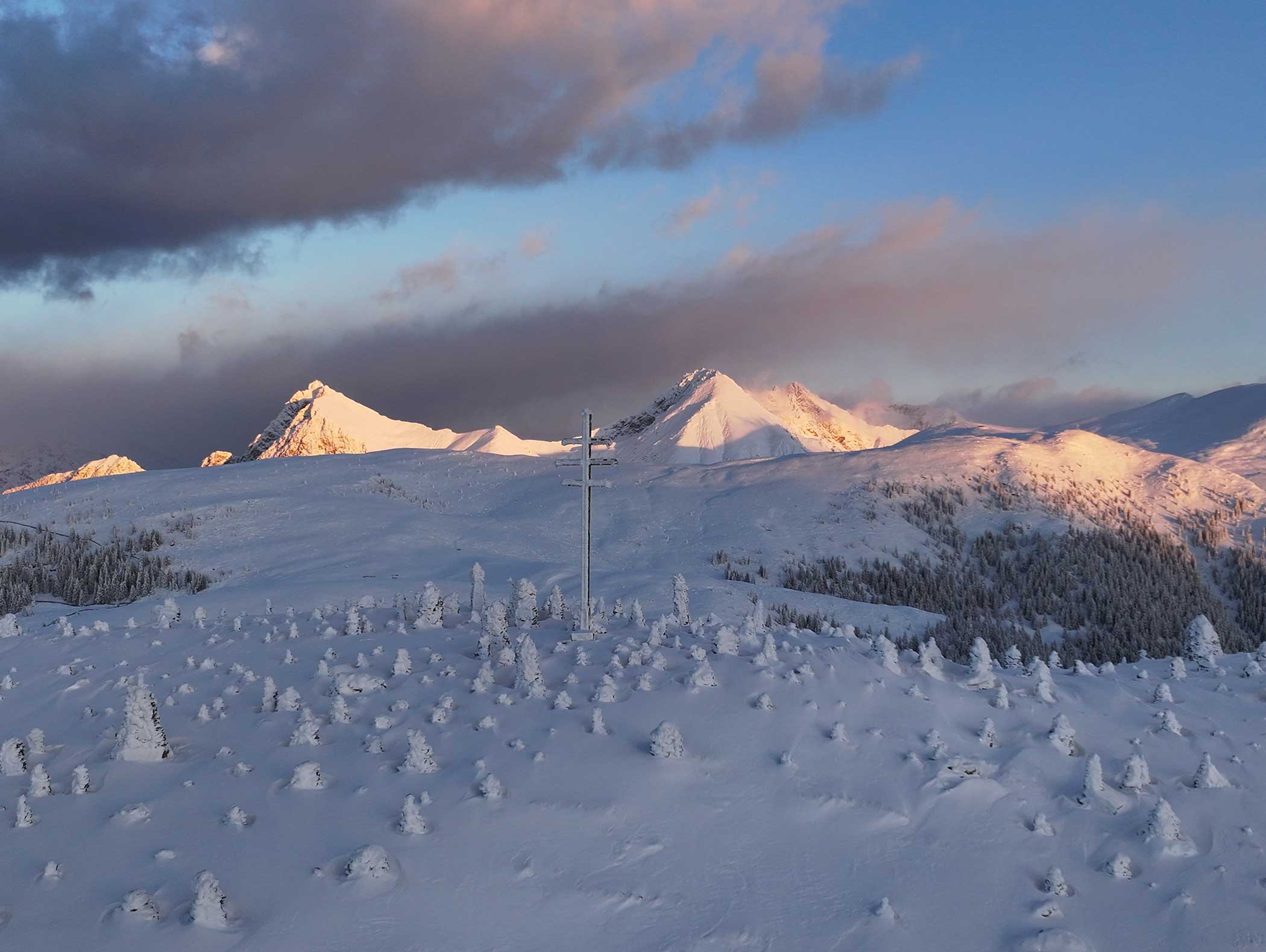 Schneebedeckte Berge bei Sonnenaufgang mit einem Kreuz auf einem Hügel und verstreuten vereisten Bäumen unter einem bewölkten Himmel. - Langfennhof