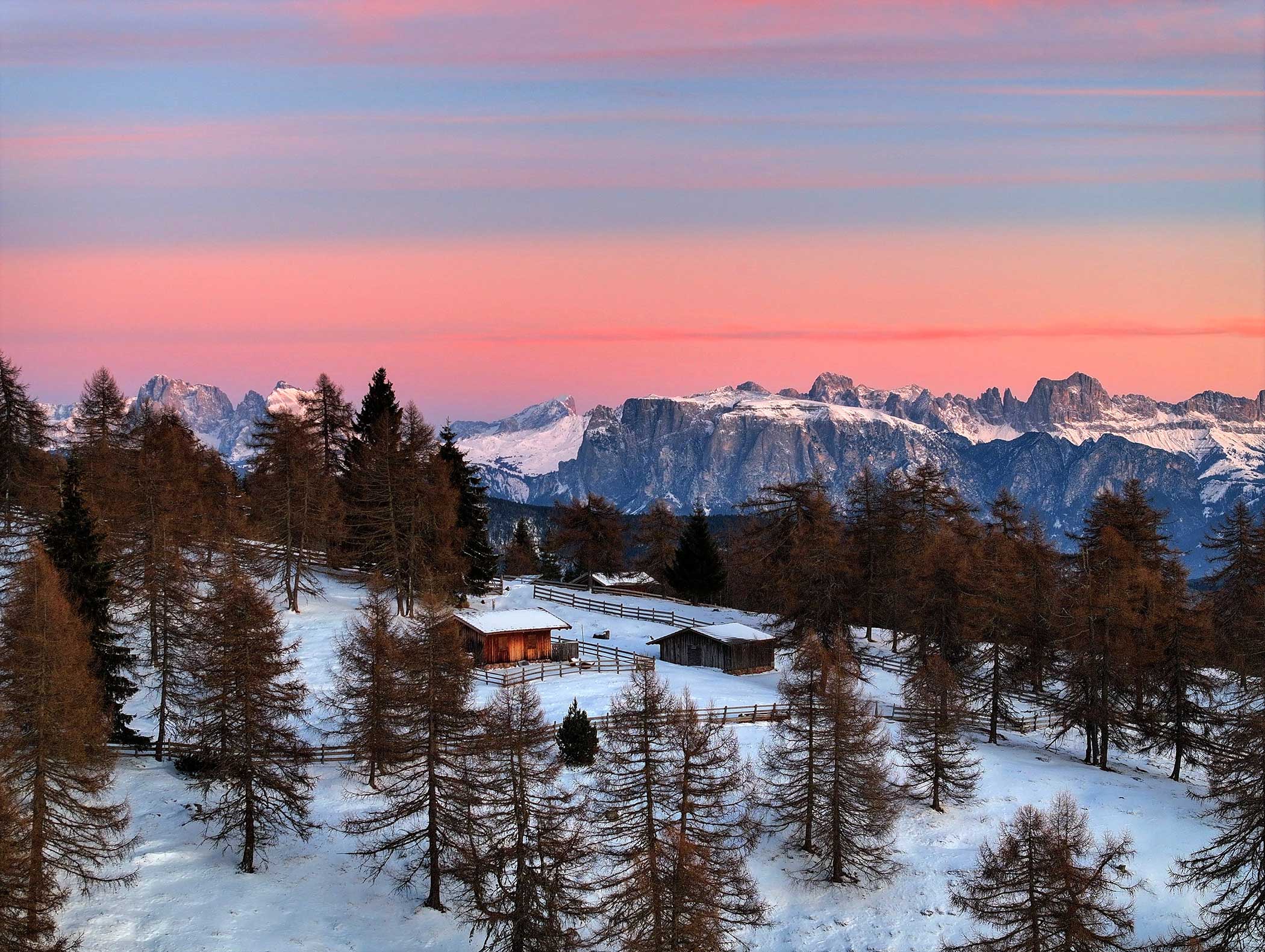 Eine verschneite Hütte zwischen Bäumen mit Bergen im Hintergrund unter einem rosafarbenen Sonnenuntergangshimmel. - Langfennhof