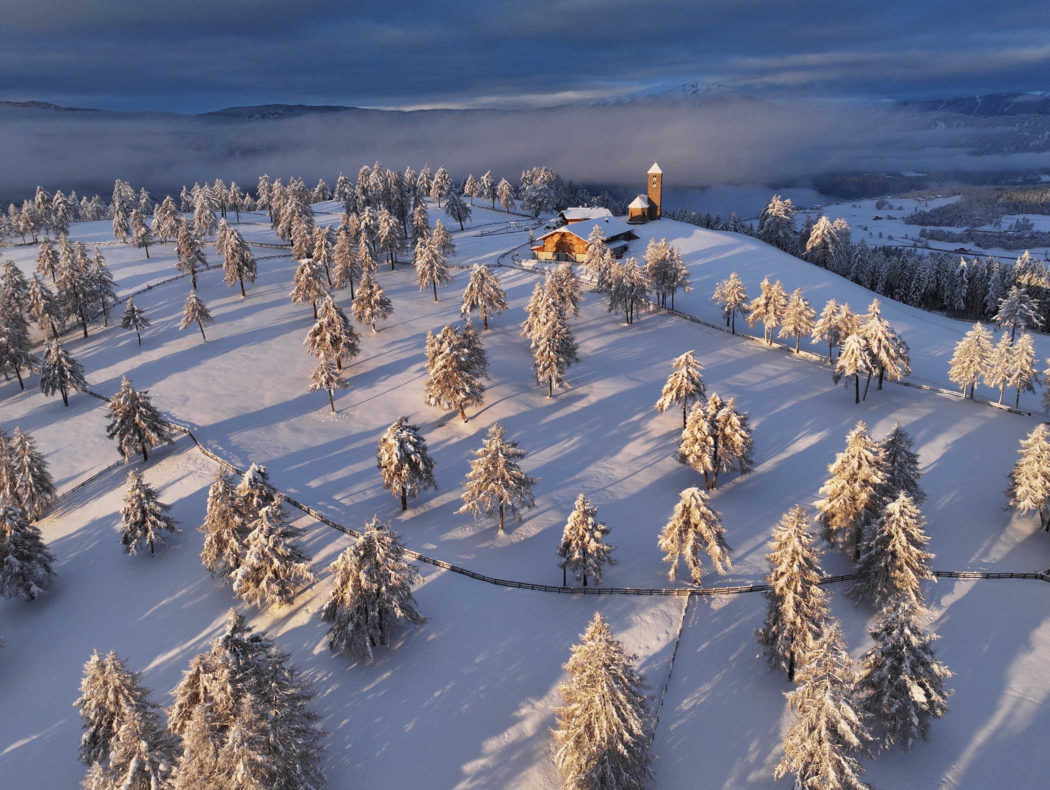 Schneebedeckte Bäume und eine Kirche auf einem Hügel in dramatischem Licht, mit kurvenreichen Straßen und Nebel in der Ferne. - Langfennhof