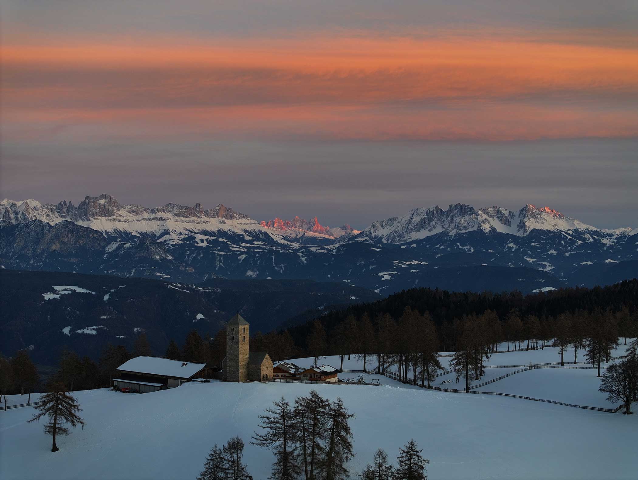 Eine schneebedeckte Landschaft mit einer kleinen Kirche, Bergen und rosa Wolken bei Sonnenuntergang. - Langfennhof