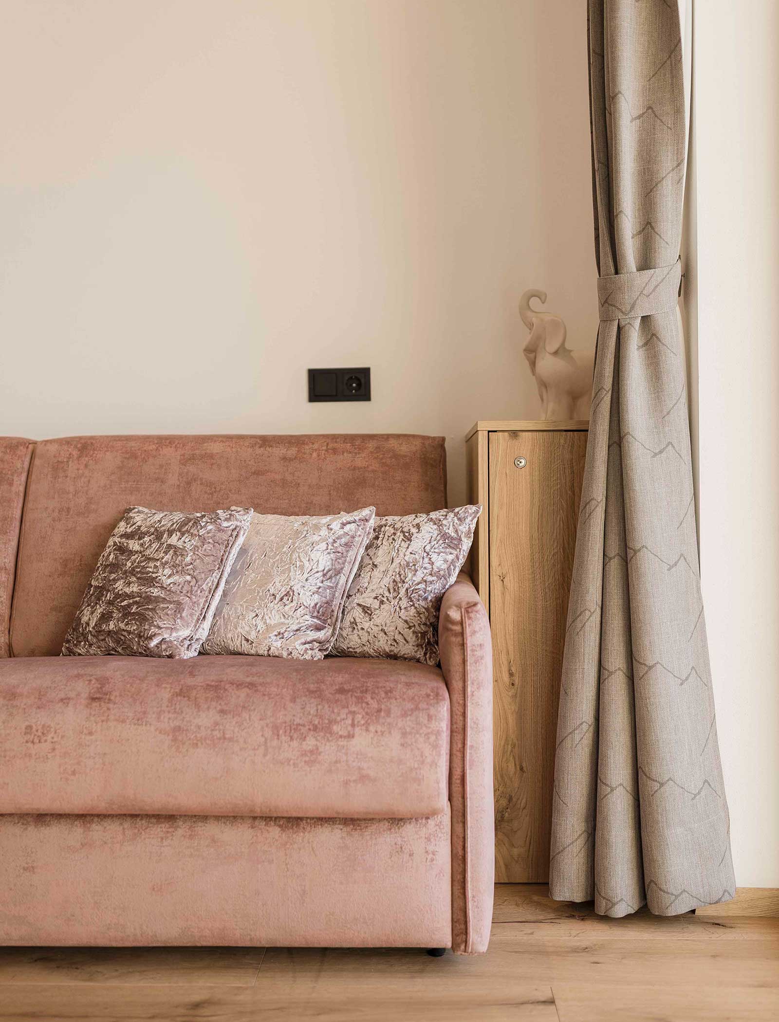 Rosa Sofa mit silbern gemusterten Kissen neben einem hölzernen Beistelltisch und beigen Vorhängen in einem gemütlichen Zimmer. - Langfennhof