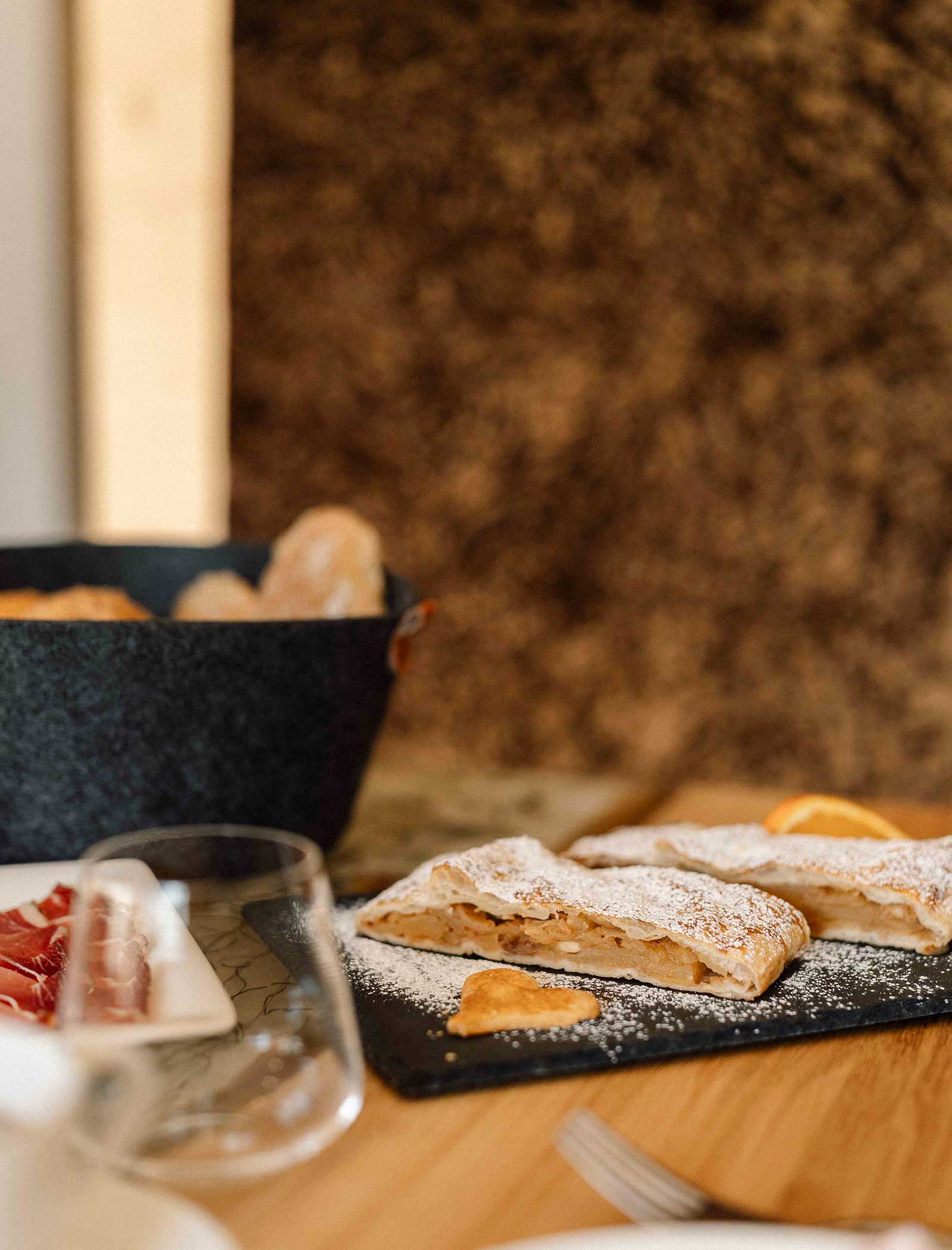 Mit Puderzucker bestäubte Apfelstrudelscheiben auf einer Schiefertafel, daneben Brot und dünn geschnittener Schinken. - Langfennhof