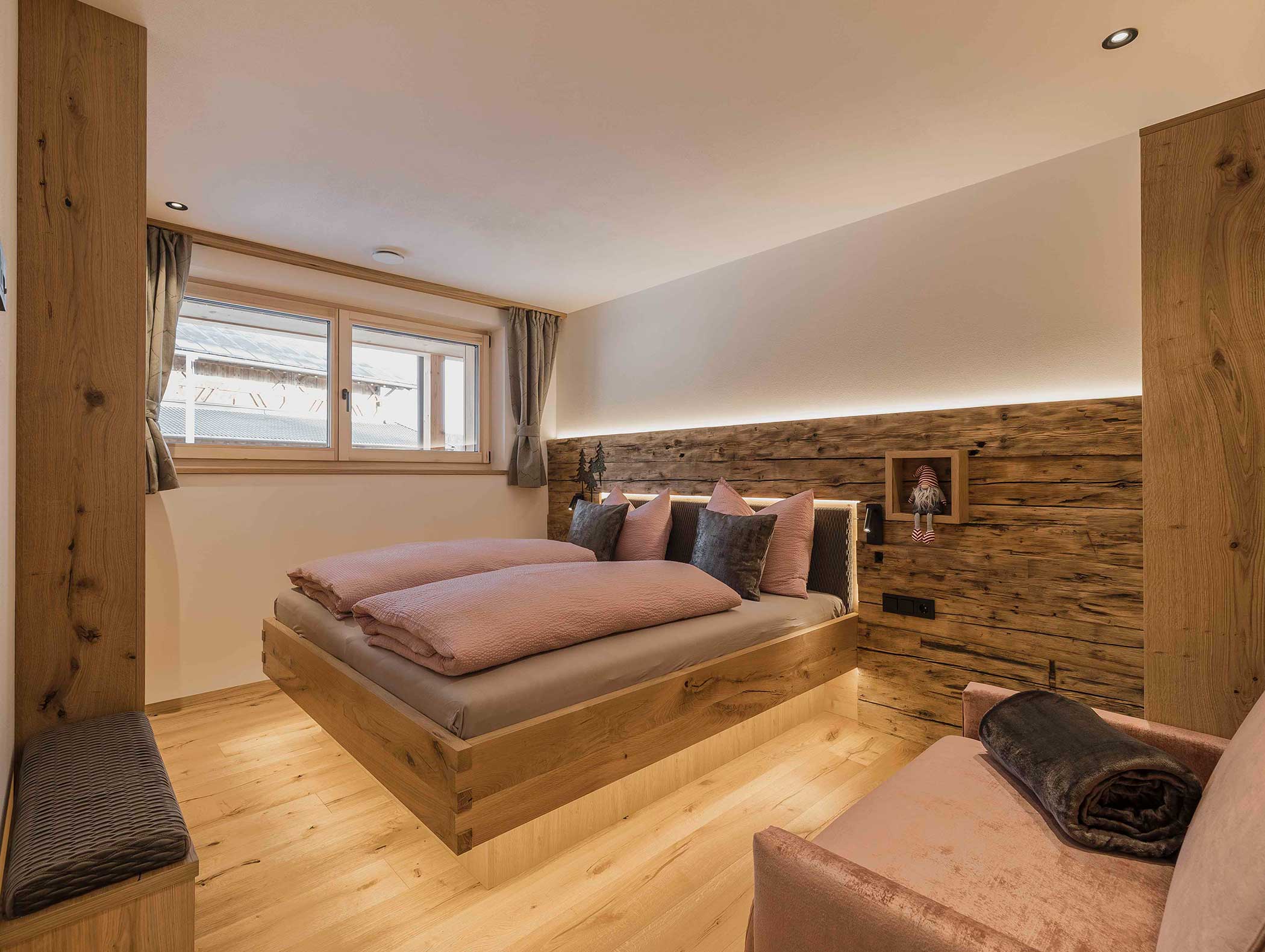 Gemütliches, modernes Schlafzimmer mit Holzakzenten, Doppelbett, sanfter Beleuchtung und einem Fenster mit grauen Vorhängen. - Langfennhof