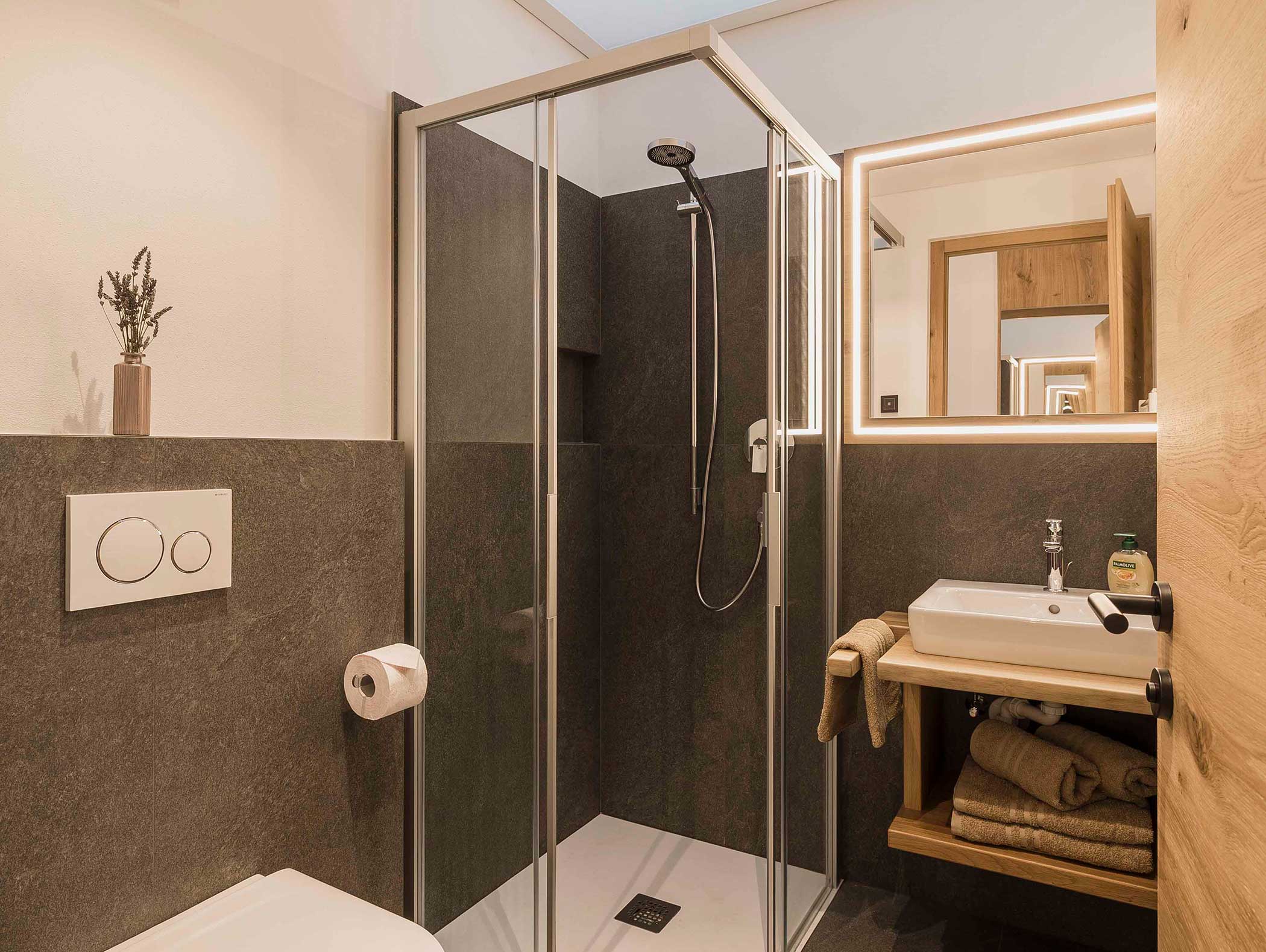 Modernes Badezimmer mit Glasdusche, wandmontierter Toilette, Waschbecken, Spiegel und ordentlich gefalteten Handtüchern auf einem Regal. - Langfennhof