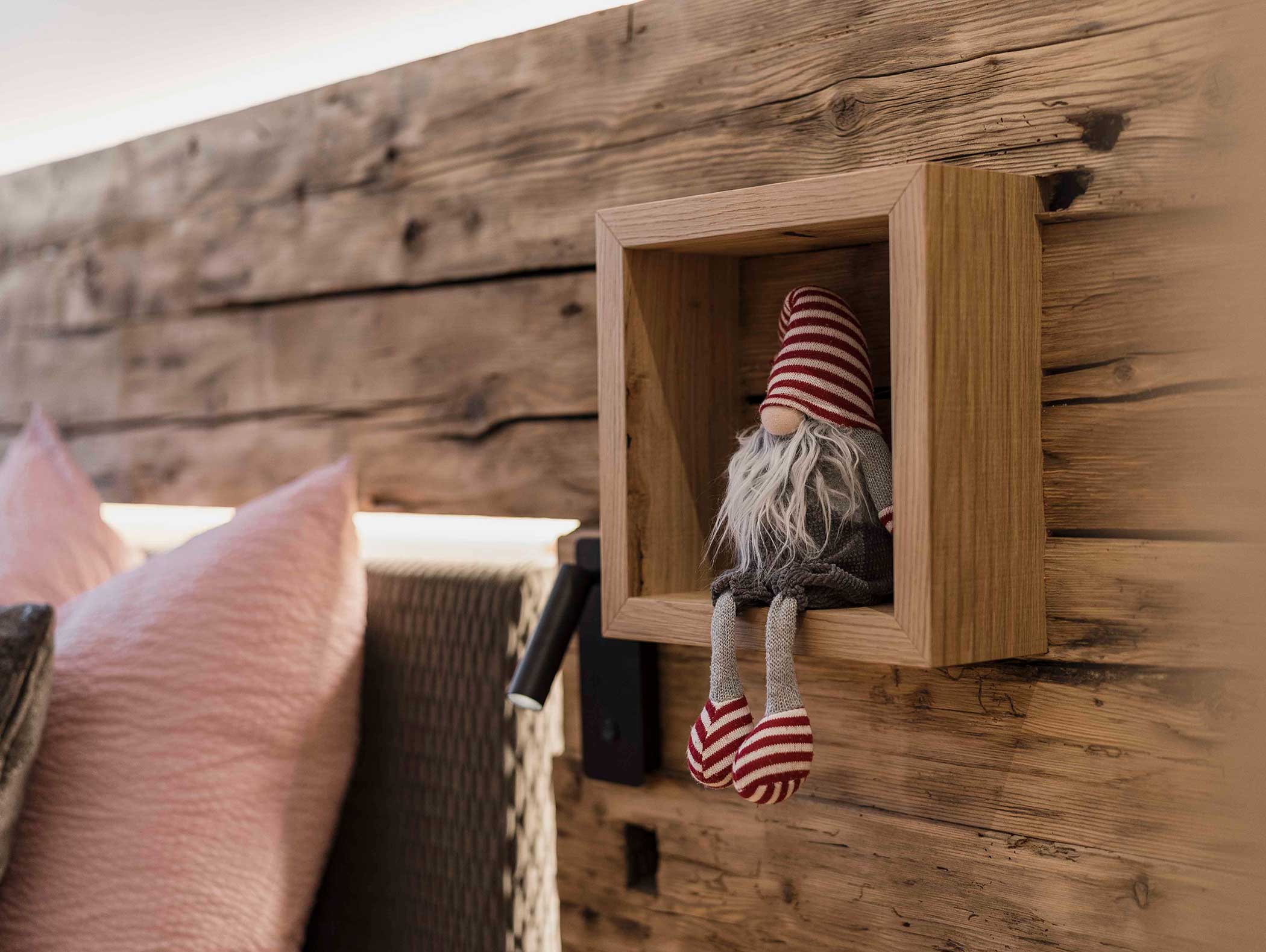 Eine kleine Zwergpuppe mit einer gestreiften Mütze sitzt in einem Holzregal an einer rustikalen Holzwand. - Langfennhof