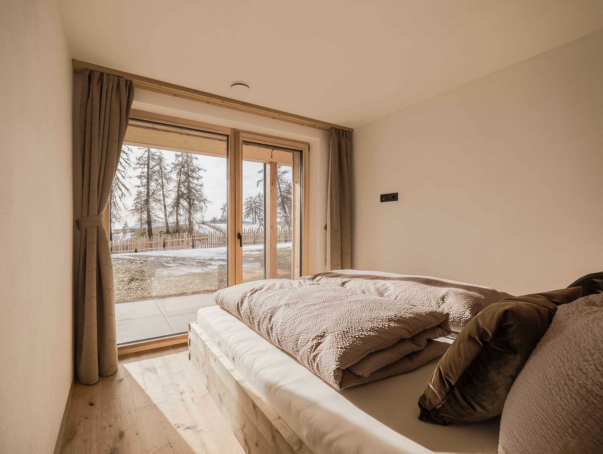 Minimalistisches Schlafzimmer mit großen Glastüren, beiger Bettwäsche und Blick auf Bäume und Schnee draußen. - Langfennhof