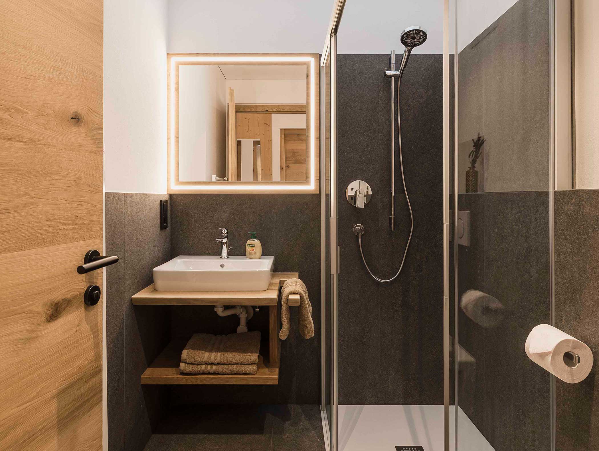 Modernes Badezimmer mit einer Glasdusche, einem Waschbecken, einem beleuchteten Spiegel und Holzakzenten. Die Handtücher sind ordentlich unter dem Waschbecken verstaut. - Langfennhof